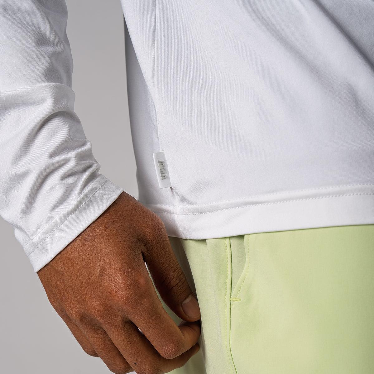 Nahaufnahme eines weißen Langarmshirts mit kleinem Markenetikett am Saum, getragen von einer Person mit hellgrüner Hose.