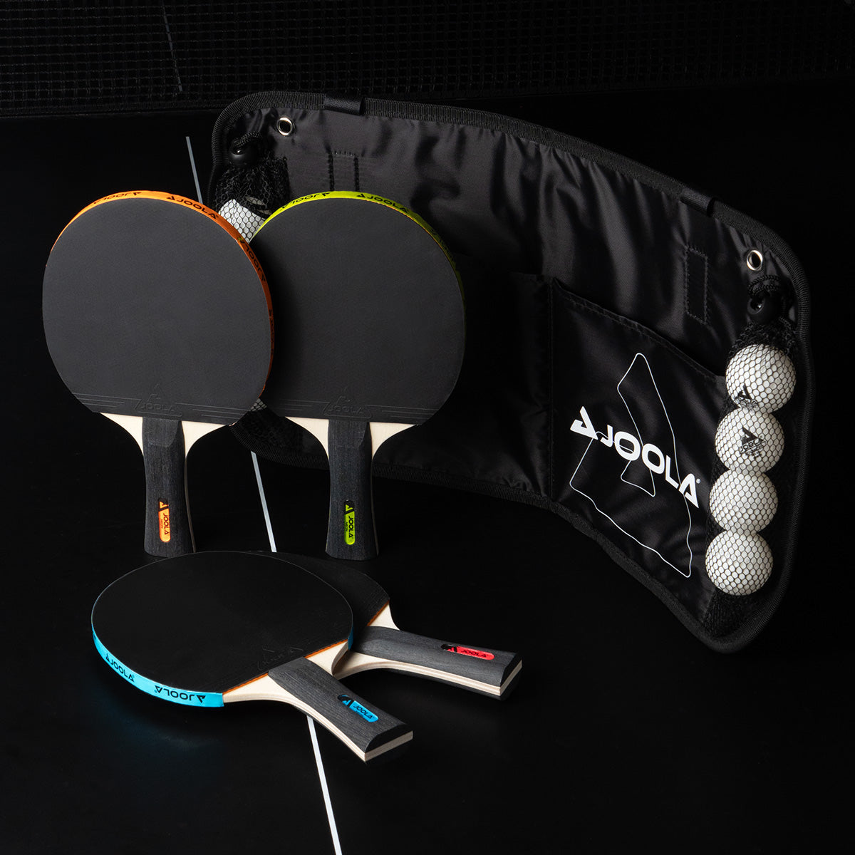 Vier Tischtennisschläger mit farbigen Griffen und ein Netz mit vier weißen Tischtennisbällen auf schwarzem Hintergrund.