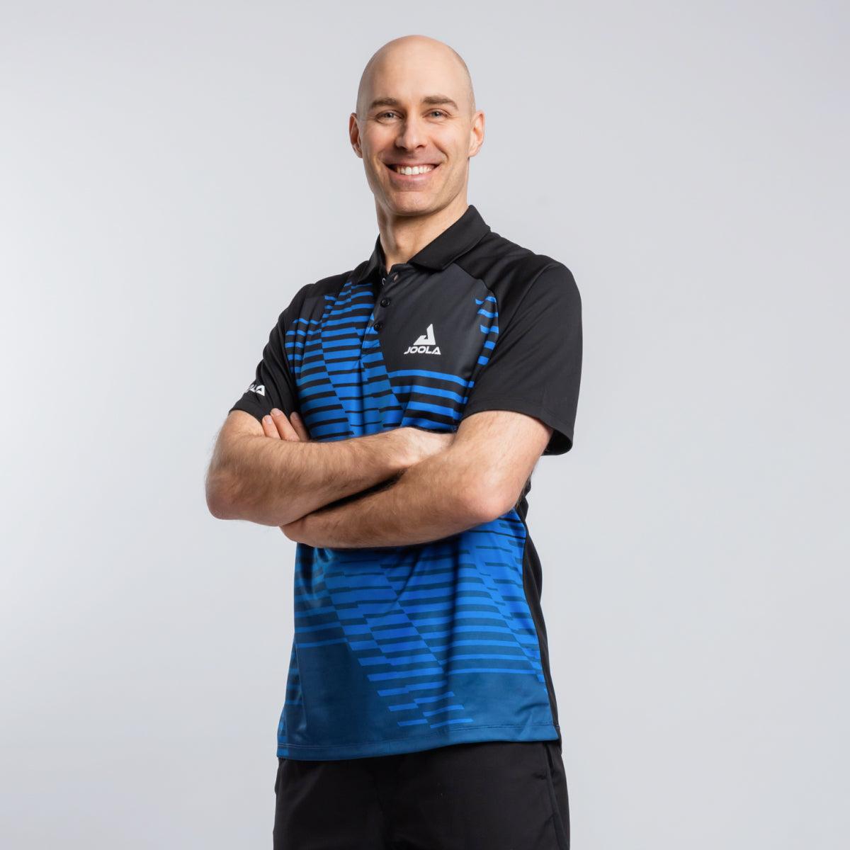 Mann trägt schwarzes und blau gestreiftes JOOLA Sport-Poloshirt, Arme verschränkt, lächelnd vor hellem Hintergrund.