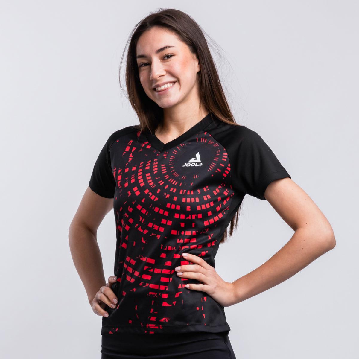Frau trägt schwarzes Sportshirt mit rotem Muster und JOOLA-Logo, lächelnd, vor weißem Hintergrund.