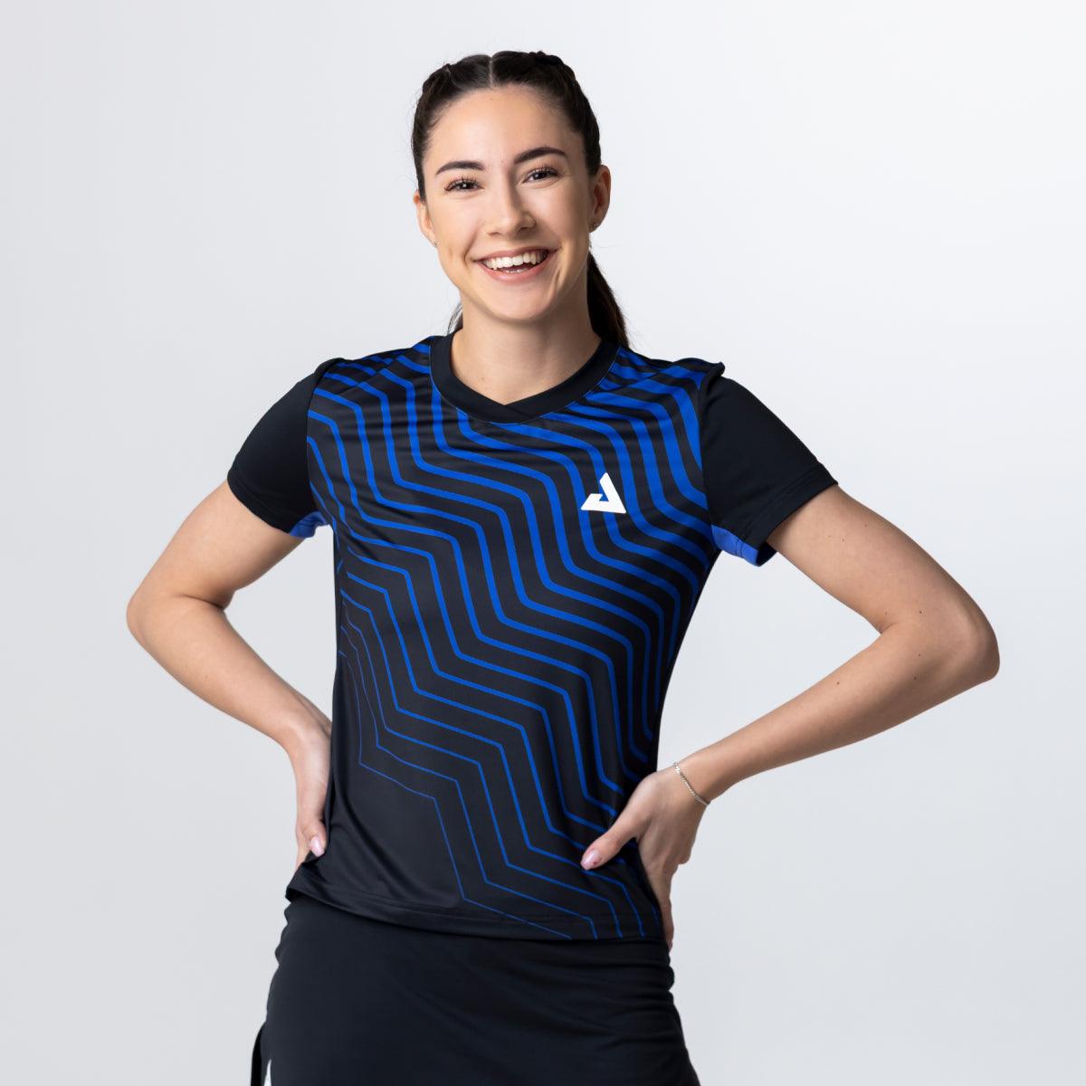 Frau trägt schwarzes Sportshirt mit blauem, wellenförmigem Muster und weißem Logo, lächelt vor weißem Hintergrund.