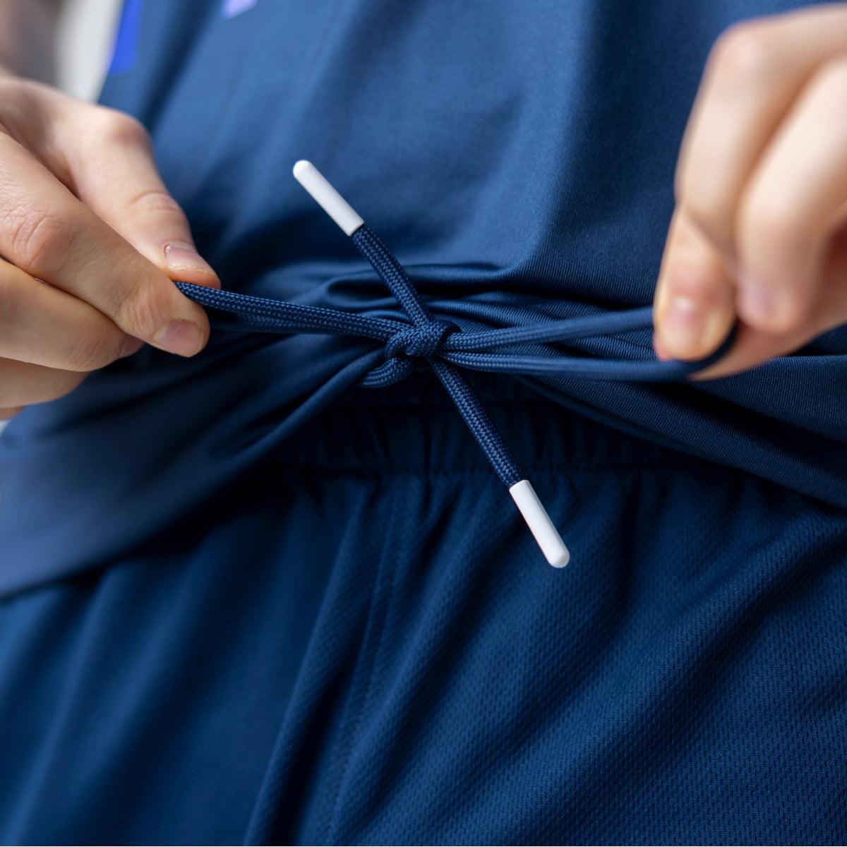 Nahaufnahme von Händen, die eine dunkelblaue Kordel an einem elastischen Bund einer dunkelblauen Sporthose zusammenziehen.
