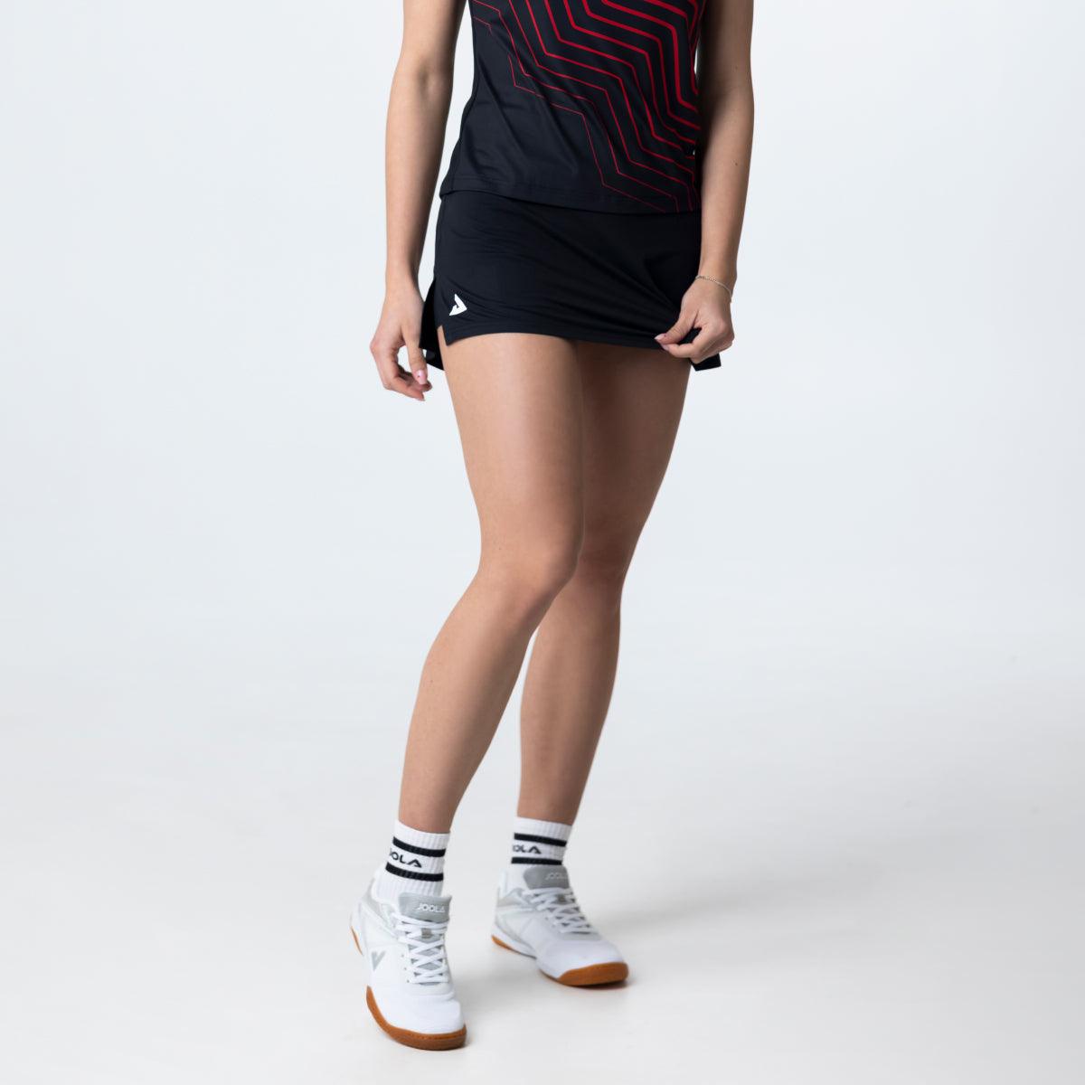 Frau trägt schwarzen Sportrock mit weißem Logo, weiße Sportsocken und weiße Sportschuhe, Vorderansicht, vor weißem Hintergrund.