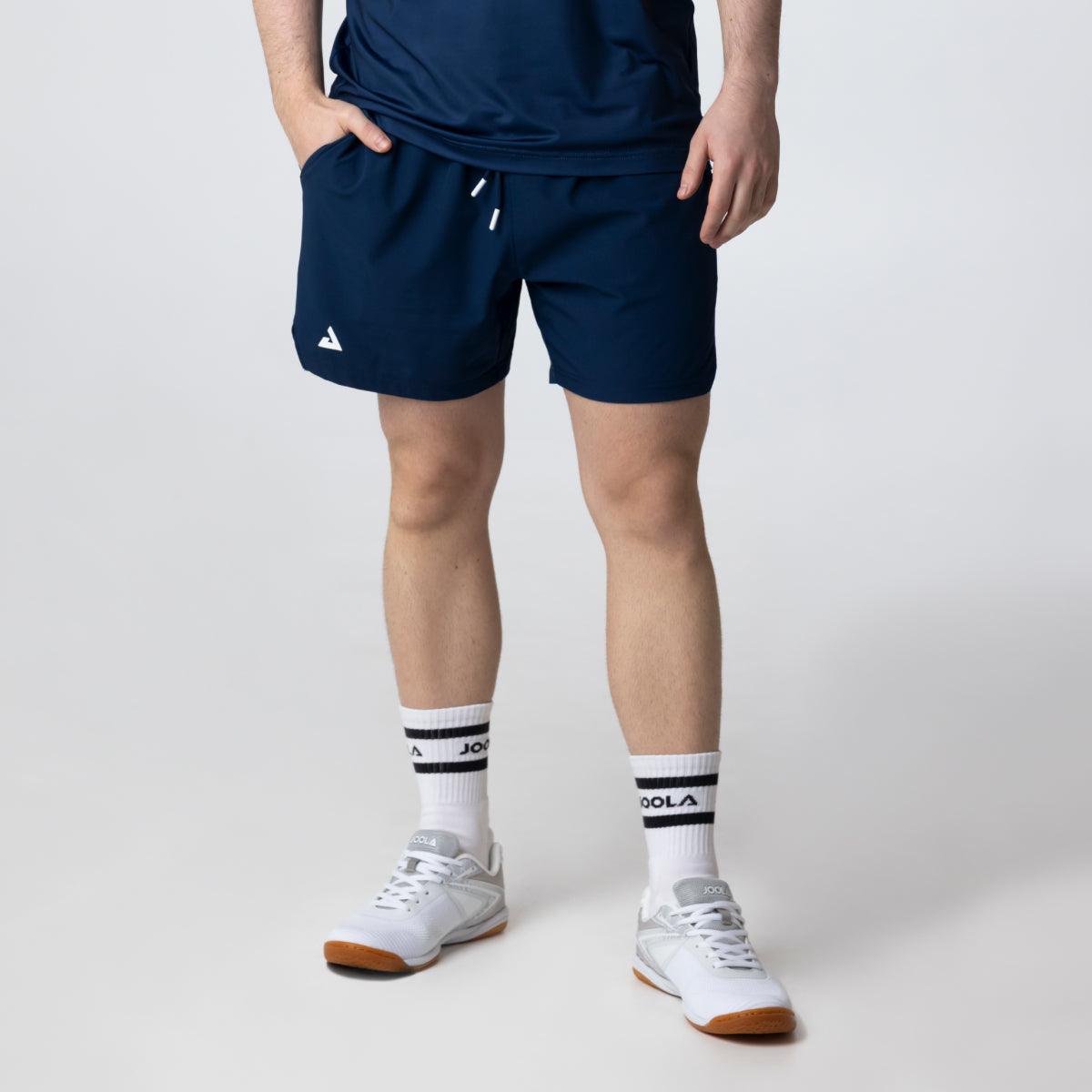 Mann trägt dunkelblaue Sportshorts mit seitlichen Taschen, weißen Sportsocken und weißen Sportschuhen vor hellem Hintergrund.