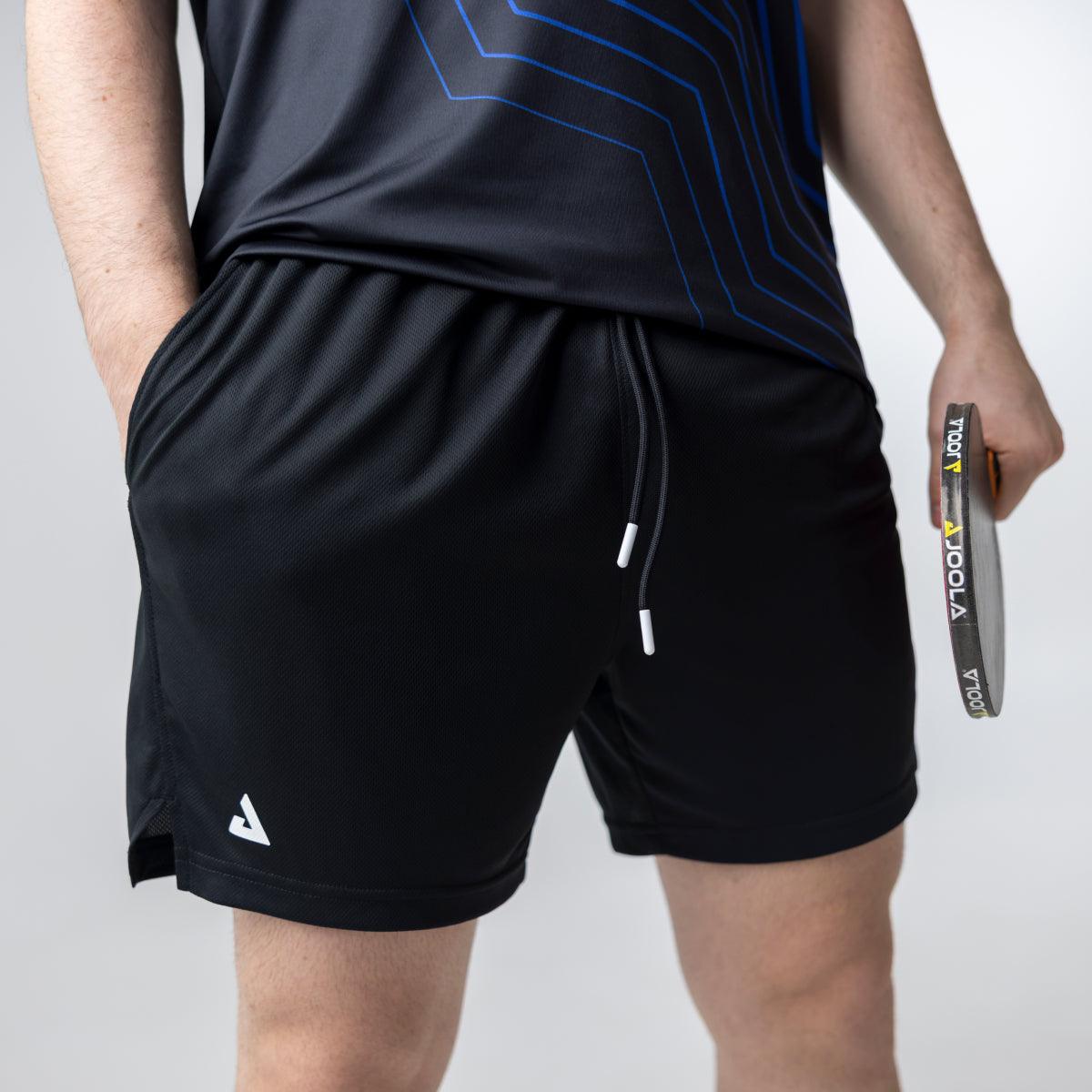 Mann trägt schwarze Sportshorts mit Kordelzug und hält einen Tischtennisschläger.